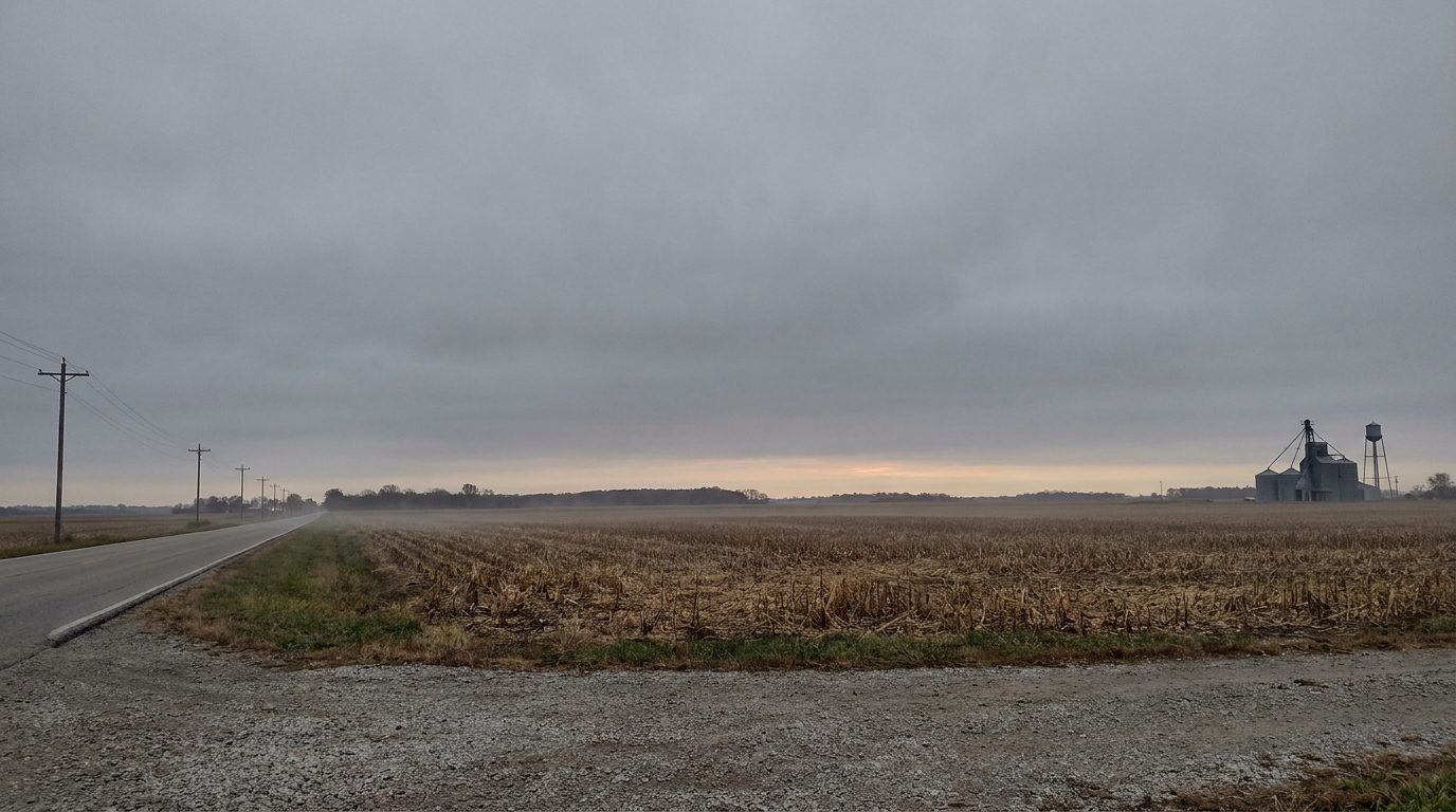 A flat central Indiana horizon at dusk, seen from the shoulder of a two-lane county road. A harvested field of stubble runs to a low treeline, a grain elevator and water tower stand on the right, utility poles run away to the left, and heavy overcast fills the sky with only a faint warm band near the horizon.